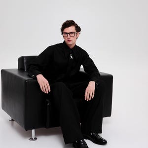 May include: A person wearing black-framed glasses sits in a black leather armchair. They are wearing a black button-down shirt with a silver pin and black pants. They are wearing black leather loafers.