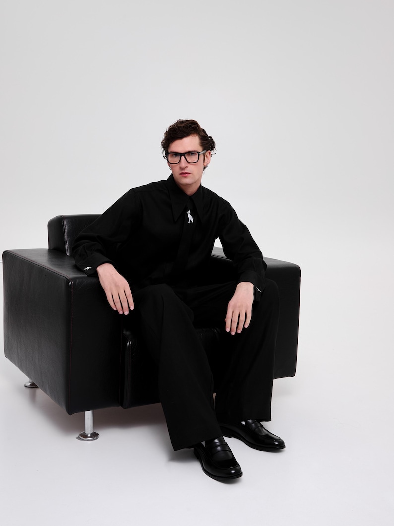 May include: A person wearing black-framed glasses sits in a black leather armchair. They are wearing a black button-down shirt with a silver pin and black pants. They are wearing black leather loafers.