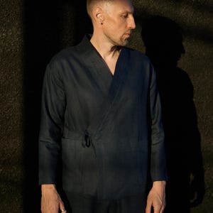May include: A dark navy blue kimono-style jacket with long sleeves and a wrap-around design. The jacket features a tie closure at the waist and a pocket on the left side. The fabric appears to be linen or a similar lightweight material. The model is wearing a matching navy blue pants.