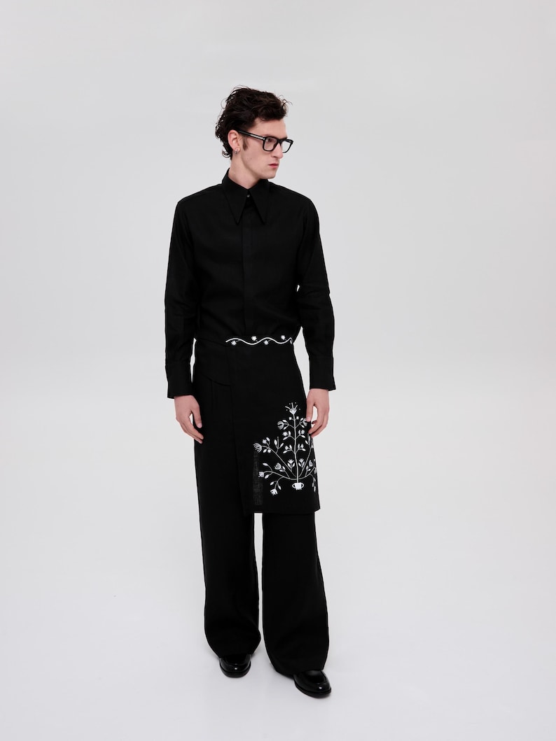 May include: A black long-sleeved shirt with a pointed collar and a white embroidered design on the front. The shirt is paired with black pants and a black belt with a white wavy line design.
