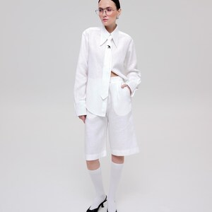 May include: A white long-sleeved button-down shirt with a pointed collar and a matching white tie featuring a small black bird emblem. The shirt is paired with white knee-length shorts, white socks, and black pointed-toe slingback heels.