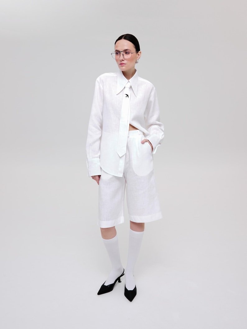 May include: A white long-sleeved button-down shirt with a pointed collar and a matching white tie featuring a small black bird emblem. The shirt is paired with white knee-length shorts, white socks, and black pointed-toe slingback heels.