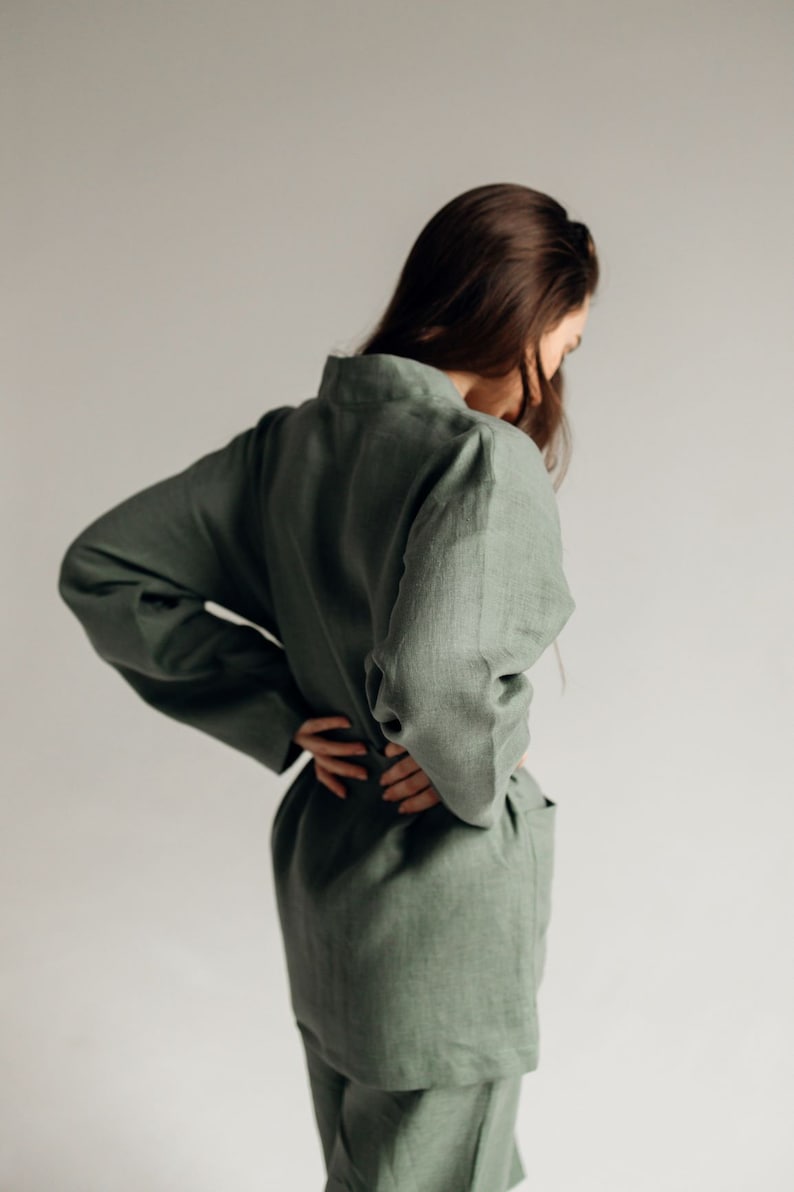 May include: A woman wearing a sage green linen jumpsuit with long sleeves and a relaxed fit.