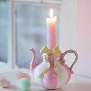 Könnte beinhalten: Ein rosa-weiß gestreifter Teekannen-Kerzenhalter mit einer brennenden rosa Kerze. Der Kerzenhalter hat einen silbernen Griff und Ausguss sowie eine gelbe Schleife. Ein Ei und eine Eierschale befinden sich im Hintergrund.