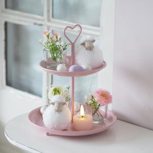 Könnte beinhalten: Ein rosa, zweistöckiger Servierständer mit herzförmigem Griff. Er zeigt kleine Glasvasen mit Blumen, flauschige weiße Schaf-Ornamente und Ostereier in Pastellfarben. Auf der unteren Ebene befindet sich eine brennende rosa Kerze.