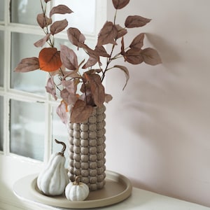 Könnte beinhalten: Eine hohe, zylindrische, beige Keramikvase mit einer texturierten, blasenartigen Oberfläche. In der Vase befinden sich mehrere Zweige mit getrockneten, bräunlich-rosa Blättern. Neben der Vase liegen zwei kleine, weiße Zierkürbisse auf einem runden, beigen Tablett.