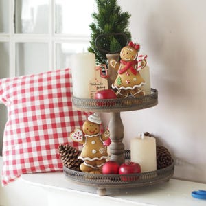 Könnte beinhalten: Ein zweistufiges dekoratives Tablett mit Weihnachtsdekorationen. Die obere Ebene enthält einen Lebkuchenmann, Kerzen und einen kleinen Weihnachtsbaum. Die untere Ebene hat einen Lebkuchenmann, rote Äpfel und Tannenzapfen.