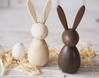 May include: Two wooden bunny figurines, one painted brown and one left natural, standing on a white surface with wood shavings.
