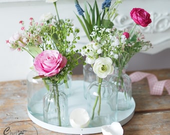 May include: A white tray with six clear glass bud vases filled with pink, white, and red flowers. The flowers are arranged in a circular pattern on the tray. There are two eggshells on the table next to the tray.