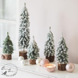 Könnte beinhalten: Eine Ansammlung von fünf künstlichen Miniatur-Tannenbäumen mit beflockten, schneebedeckten Nadeln, die jeweils auf einem natürlichen Holzscheibenfuß stehen. Das Arrangement enthält glatte, runde Kugeln in Weiß und Zartrosa sowie einen kleinen Kerzenhalter.