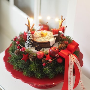 Könnte beinhalten: Ein weihnachtliches Tischgesteck auf einem roten, gewellten Ständer mit einem Miniatur-Weihnachtsmann in einer schaumgefüllten Holzwanne, einer gelben Quietscheente, einem kleinen Tannenbaum und zwei braunen Ästen mit einer Lichterkette.