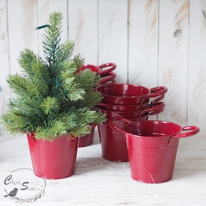 Könnte beinhalten: Ein kleiner künstlicher Weihnachtsbaum in einem roten Metalleimer. Der Baum ist von mehreren weiteren roten Metalleimern umgeben, einige übereinander gestapelt.