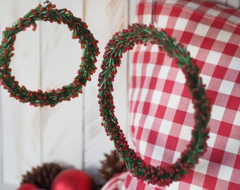 May include: Two decorative wreaths with green and red beaded details hang in front of a red and white checkered pillow. The wreaths are circular and appear to be handmade. Red Christmas ornaments and pinecones are visible below.