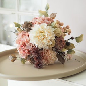 Könnte beinhalten: Ein Blumenarrangement mit einer zentralen cremefarbenen Dahlie, umgeben von altrosa Hortensien, kleineren strukturierten Blüten, getrocknet wirkendem Blattwerk und Clustern aus kleinen runden Beeren, angeordnet zu einem runden Strauß.