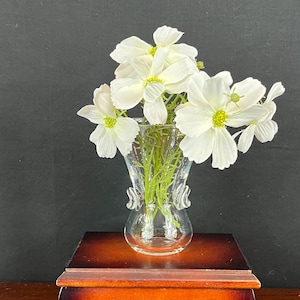 May include: A small, clear glass vase with white flowers and green stems sits atop a wooden box with a curved design. The box has a dark brown finish with lighter brown accents. The background is a dark gray.