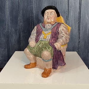 May include: A ceramic figurine of a seated man in historical attire. The figure wears a black hat, purple vest, gold necklace, and green shorts. The figurine is seated on a decorative chair and has a detailed, hand-painted appearance.