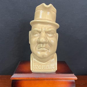 May include: A tan ceramic bust of W.C. Fields wearing a hat, with the name "W.C. FIELDS" on the base. The bust sits on a small, two-tone wooden box with a curved design. The background is dark.
