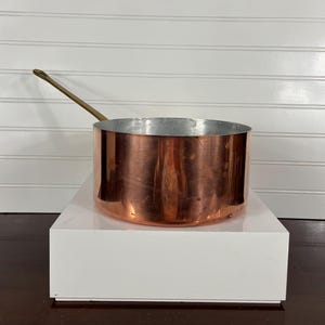 May include: A copper saucepan with a long, brass handle. The pot has a shiny, reflective surface and a silver interior. The cookware is cylindrical and sits on a white platform against a white-paneled wall.