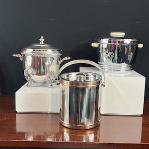 May include: Three silver ice buckets, one with a lid and two without. The ice bucket on the left has a decorative design and sits on a white pedestal. The ice bucket on the right has a copper band around the top. All three ice buckets are on a wooden surface.