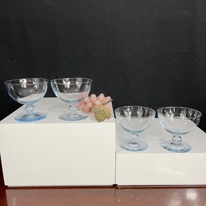 May include: Four clear glass dessert bowls with a light blue tint and a rounded base. The bowls are arranged in two rows on a white surface.