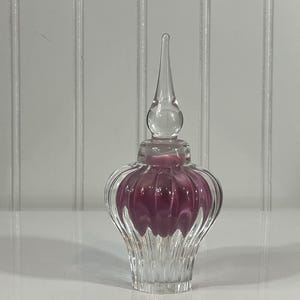 May include: A clear glass perfume bottle with a decorative, fluted design. The bottle has a bulbous, ribbed body with a deep purple interior and a clear, pointed stopper. The bottle sits on a white surface.