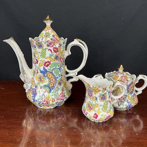 May include: A vintage porcelain tea set with a floral pattern in shades of red, blue, yellow, and green. The set includes a teapot, creamer, and sugar bowl, all with gold accents.