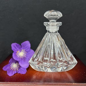May include: A clear glass bottle with a faceted design and a crystal stopper. The bottle is sitting on a wooden surface with two purple flowers.