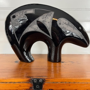 May include: A glossy black ceramic sculpture of a stylized bear. The bear has a rounded body with a curved back and short legs. The bear's body is decorated with a white and gray abstract design. The sculpture is displayed on a wooden surface.