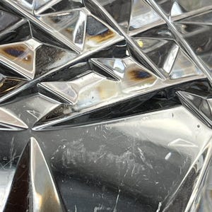Puede incluir: Primer plano de un objeto de vidrio transparente con un patr&oacute;n geom&eacute;trico. El vidrio tiene una superficie reflectante y muestra signos de desgaste.