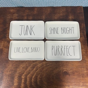 May include: Four white ceramic trays with black handwritten text. The trays read "JUNK", "SHINE BRIGHT", "LIVE, LOVE, BARK!" and "PURRFECT".