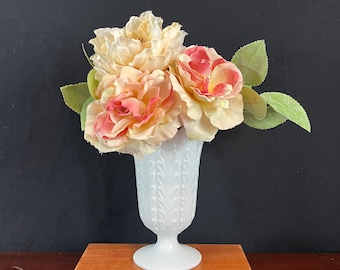 May include: A white milk glass vase with a textured design holds a bouquet of peach and pink artificial roses.