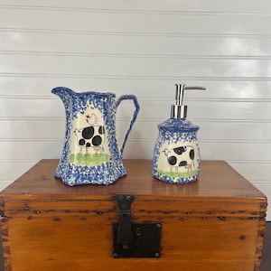 May include: A ceramic pitcher and soap dispenser set. The pitcher has a handle and a blue floral pattern with a cow and chicken illustration. The soap dispenser has a silver pump and a matching cow illustration.