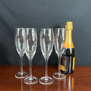May include: Four clear champagne flutes with gold dot accents and a champagne bottle with a gold foil top. The flutes are arranged in a row on a wooden surface, with the bottle to the right.