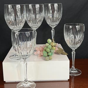 May include: Set of five clear glass wine glasses with a cut crystal design. The glasses are arranged on a white surface with a bunch of green and pink grapes in the centre.