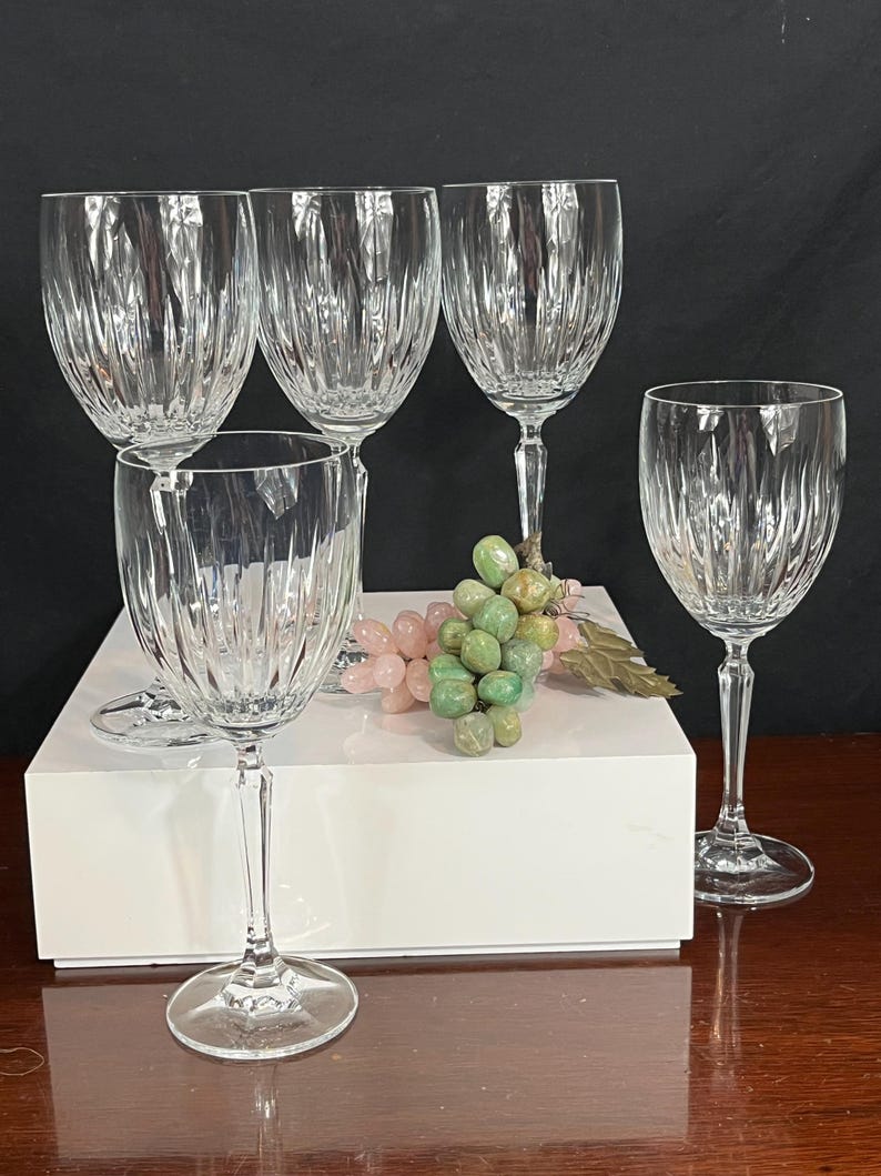 May include: Set of five clear glass wine glasses with a cut crystal design. The glasses are arranged on a white surface with a bunch of green and pink grapes in the centre.