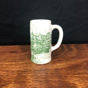 May include: A white ceramic mug with a green image of Caernarfon Castle. The mug has a handle and is on a wooden surface.