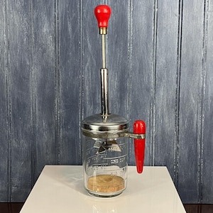 May include: A vintage glass butter churn with a red plastic handle and a red plastic knob on the top. The churn has a glass jar with measurements marked on the side.