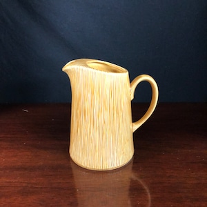 May include: A yellow ceramic pitcher with a textured, wood-grain design. The pitcher has a wide mouth and a handle.