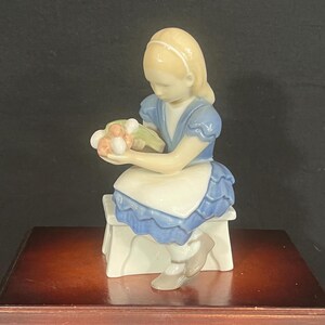 May include: A porcelain figurine of a young girl in a blue dress and white apron, seated on a small bench. She holds a bouquet of flowers. The figurine has a light skin tone and blonde hair. The base is white.