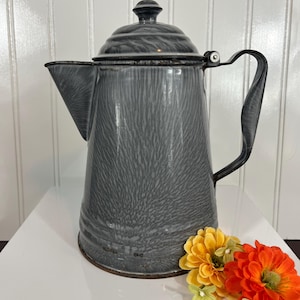 May include: A gray enamelware coffee pot with a lid and handle. The pot has a speckled pattern and a spout. The handle is black. The pot is sitting on a white surface with orange and yellow flowers.