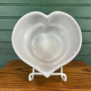 Può includere: Una ciotola a forma di cuore in ceramica grigio chiaro. La ciotola è sostenuta da un supporto in metallo bianco. La ciotola ha una finitura liscia e lucida ed è vuota. Lo sfondo è un muro di legno verde.