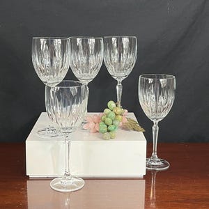 May include: Set of four clear glass wine glasses with a cut glass design. The glasses are arranged on a white surface with a bunch of green and pink grapes in the centre.