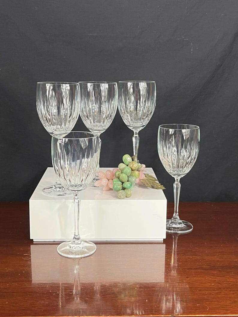 May include: Set of four clear glass wine glasses with a cut glass design. The glasses are arranged on a white surface with a bunch of green and pink grapes in the centre.