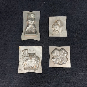 May include: Four vintage metal molds with embossed designs. One mold features a baseball player, another an indigenous person's profile, a camel, and a four-leaf clover. The molds are silver-colored and appear to be made of aluminum.