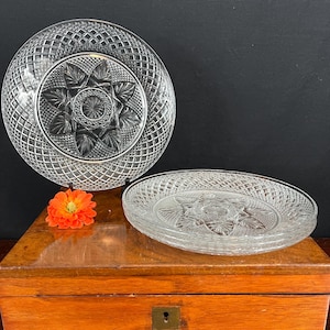 May include: A set of clear pressed glass plates featuring a central starburst pattern surrounded by a diamond lattice design. The plates have a scalloped edge and are arranged with one standing upright and two stacked horizontally.