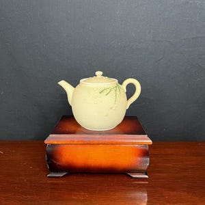 May include: A small, light yellow teapot with a green floral design sits on a wooden box with a dark brown finish.
