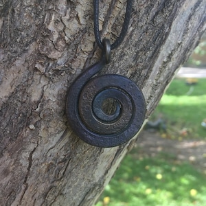 May include: A dark brown metal spiral pendant necklace on a black cord. The pendant is hanging from a tree branch.