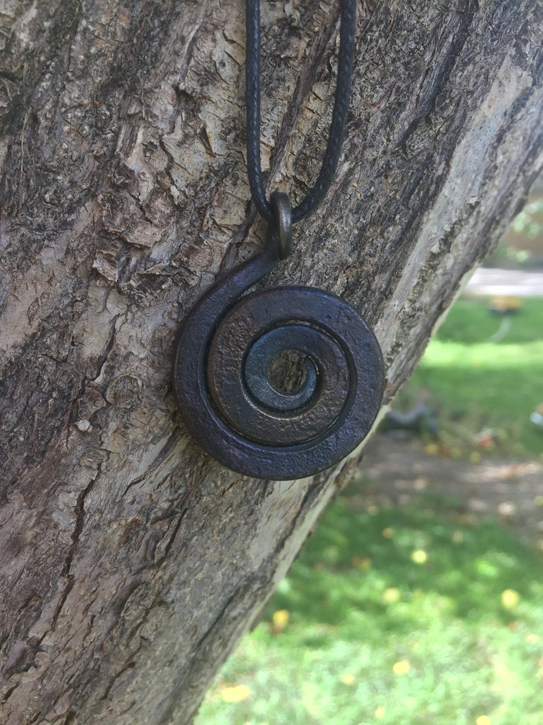 May include: A dark brown metal spiral pendant necklace on a black cord. The pendant is hanging from a tree branch.