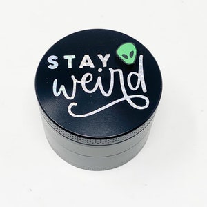以下が含まれることがあります： 緑色のエイリアンのステッカーと白い文字で「STAY weird」と書かれた黒い金属製のグラインダー