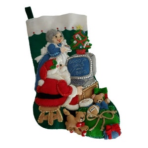 Może przedstawiać: Ręcznie robiona pończocha świąteczna z zielonym tłem i białą górą. Święty Mikołaj jest przedstawiony w kolorze czerwono-białym, siedząc przy komputerze z napisem "GOOD GIRLS AND BOYS". Inne dekoracje to choinka, misie i prezenty.